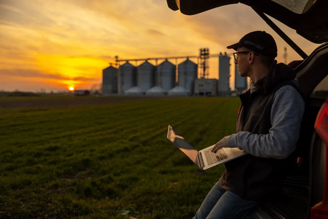 A farmer checking a laptop at sunset near grain silos
